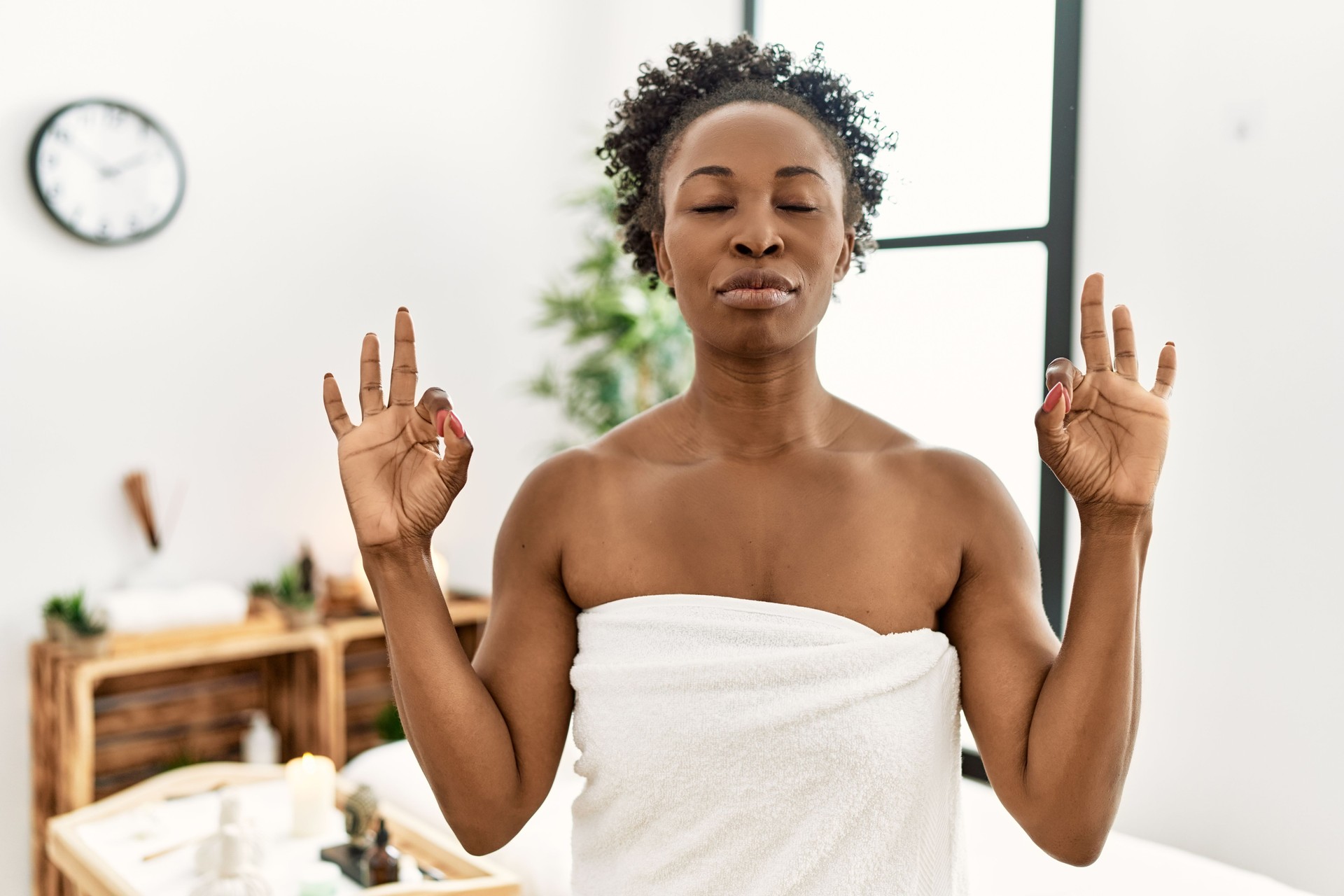 Young african american woman wearing towel standing at beauty center relax and smiling with eyes closed doing meditation gesture with fingers. yoga concept.