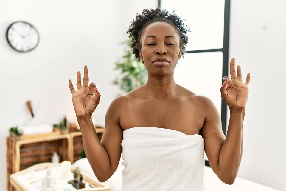 Young african american woman wearing towel standing at beauty center relax and smiling with eyes closed doing meditation gesture with fingers. yoga concept.